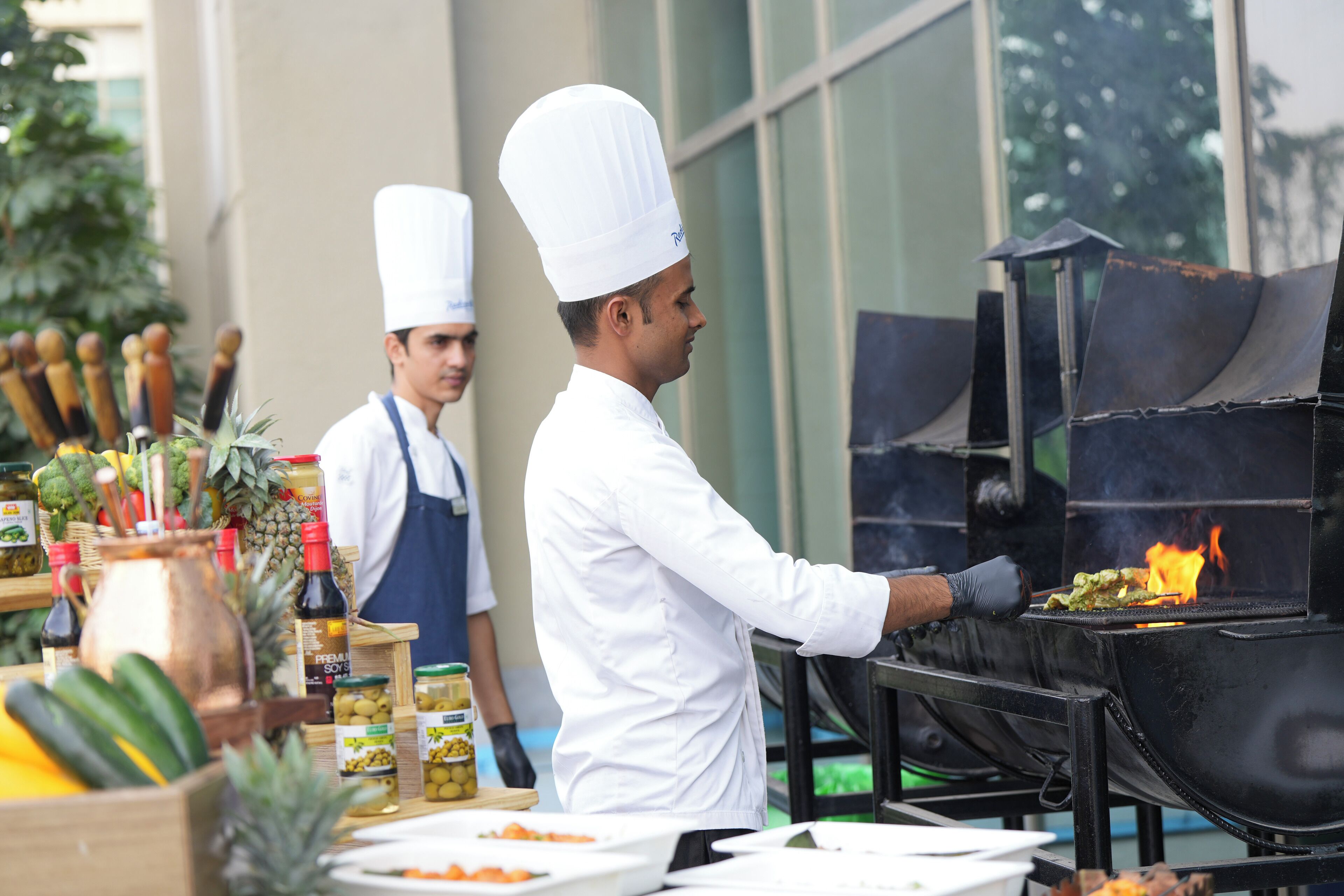 Ein Koch bereitet Essen im Freien auf einem Grill zu, verschiedene Gewürze sind sichtbar.