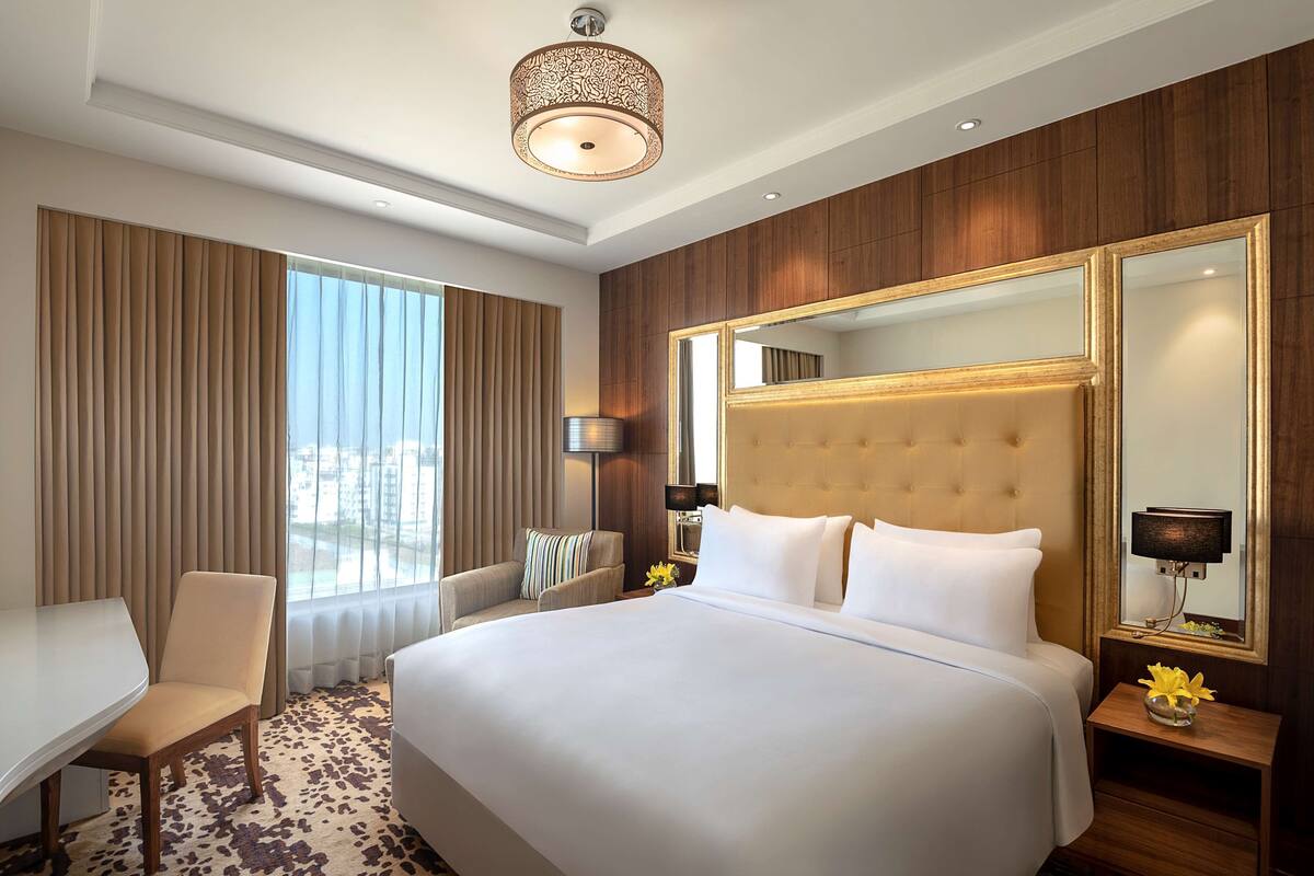 Habitación de hotel con una cama grande, un escritorio, una silla, un espejo amplio y vistas a la ciudad por la ventana.
