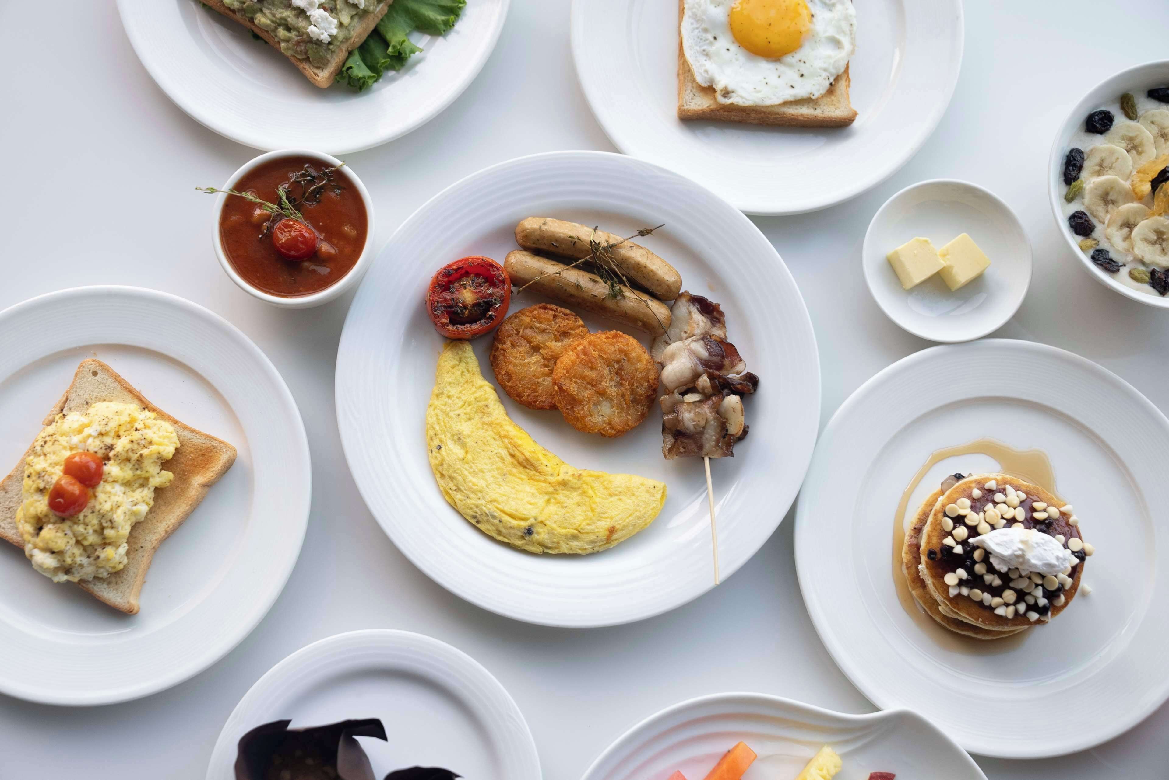 A variety of breakfast dishes on white plates, including scrambled eggs, sausages, bacon, and pancakes.