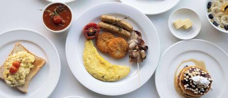 A variety of breakfast dishes on white plates, including scrambled eggs, sausages, bacon, and pancakes.