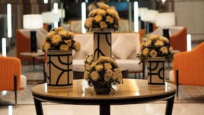 A modern hotel lobby with a round table in the foreground, featuring three vases with white flowers. A large, geometric chandelier hangs from the ceiling, and the room has a contemporary design with orange chairs and a large window in the background. - Radisson Blu Pune Hinjawadi (Paud)