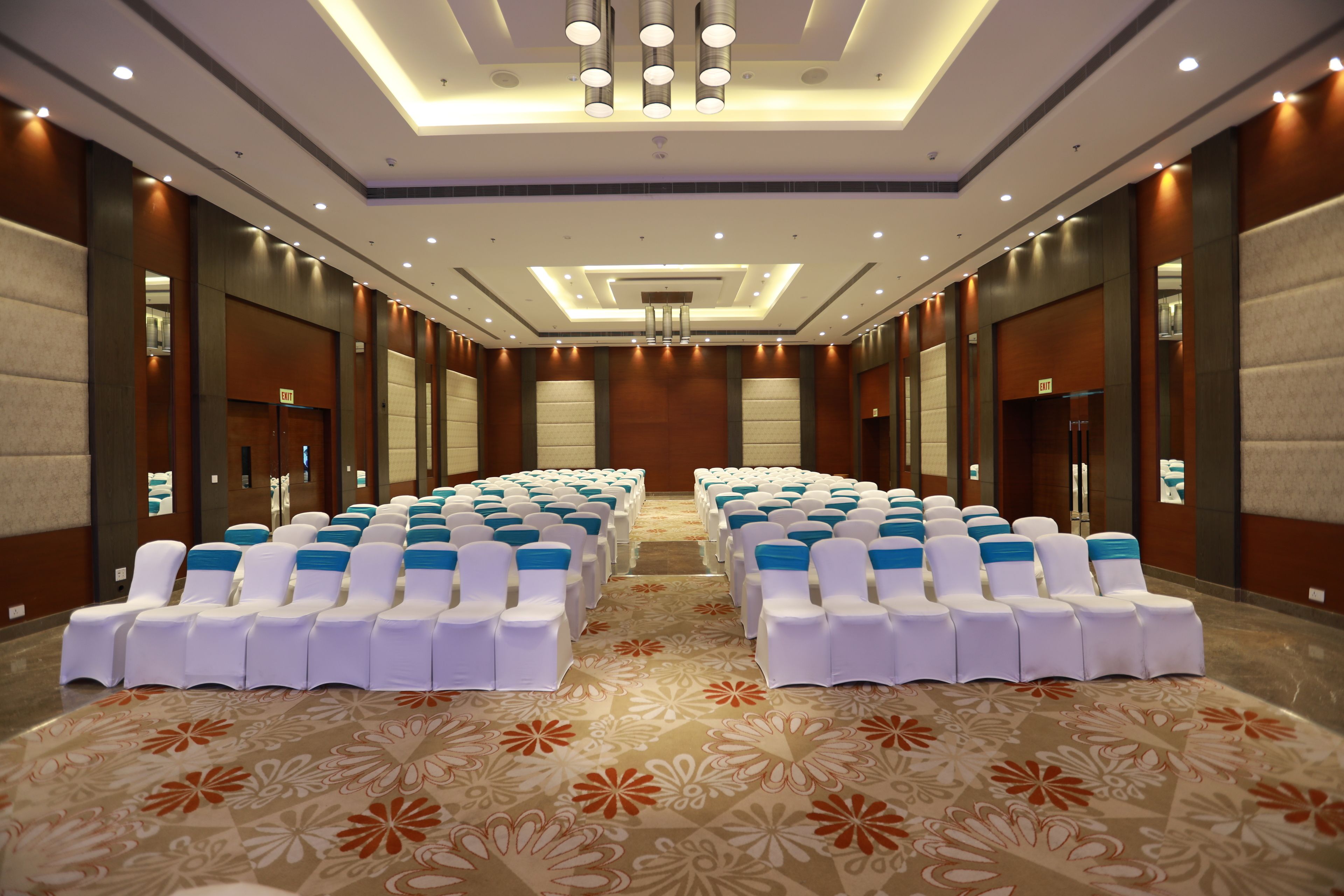 A hotel conference room with rows of white chairs with blue sashes, a patterned carpet, and a modern ceiling design.