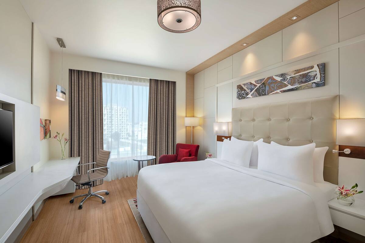 Habitación de hotel con una cama grande, un escritorio con televisor de pantalla plana, una silla roja, una mesita pequeña y un ventanal con cortinas.