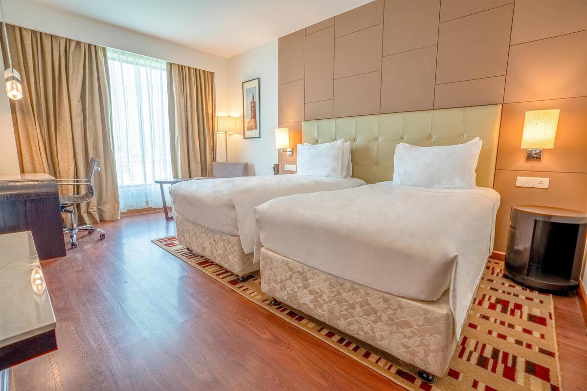 Habitación de hotel con dos camas, un escritorio, una silla, una lámpara y vistas al exterior.