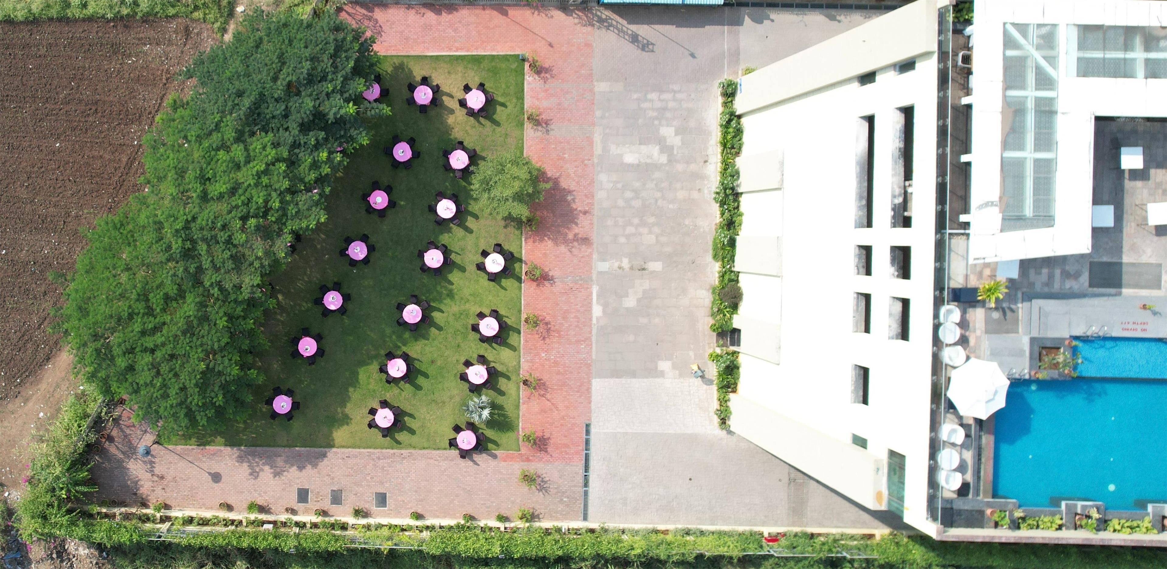 Vista aèria d'un hotel amb una piscina al terrat, un jardí paisatjistic amb taules i cadires, i una façana d'edifici moderna.