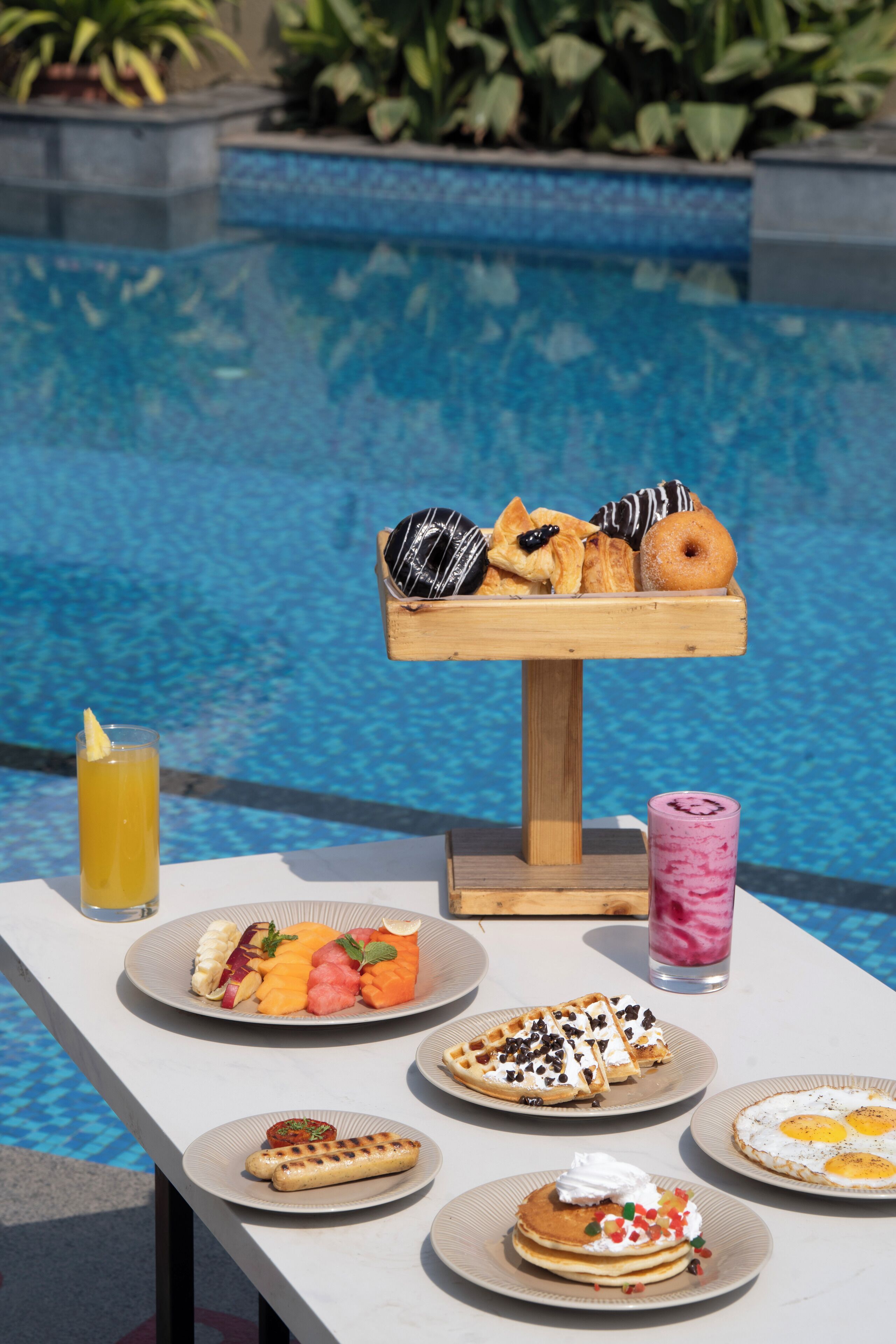 Desayuno junto a la piscina con diversos pasteles, gofres y una bandeja de fruta, además de bebidas.