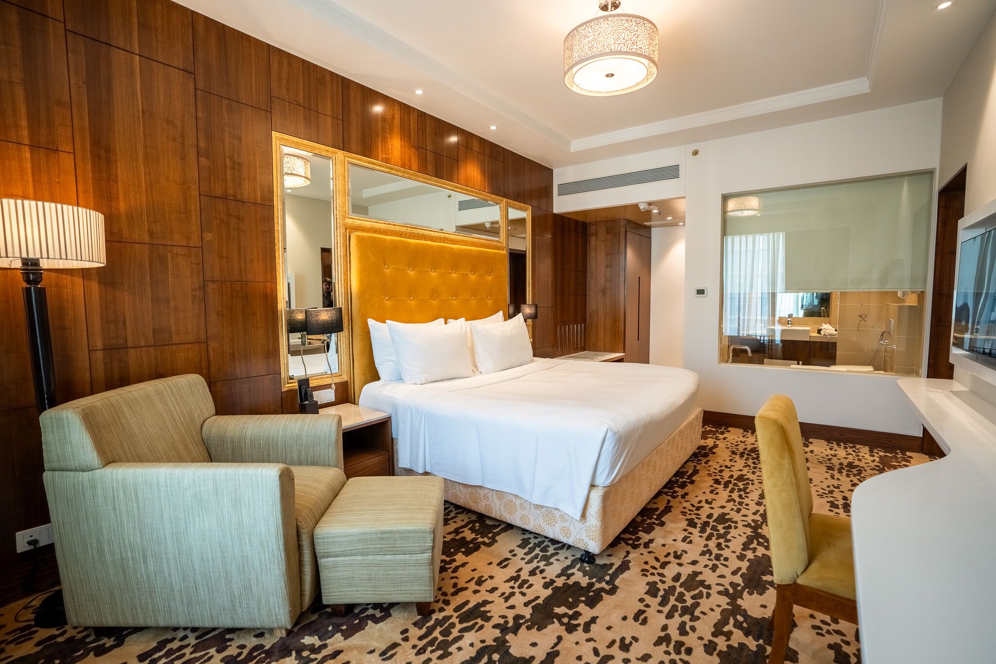 A hotel room with a large bed, a sitting area with a chair and ottoman, a mirror, and a bathroom visible through a glass door.