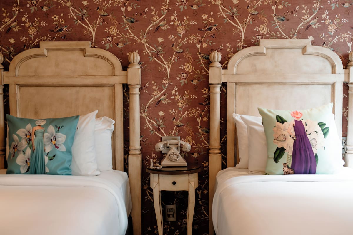 A room with two beds, each with floral-patterned pillows, a small table with a telephone, and a floral wallpaper design.