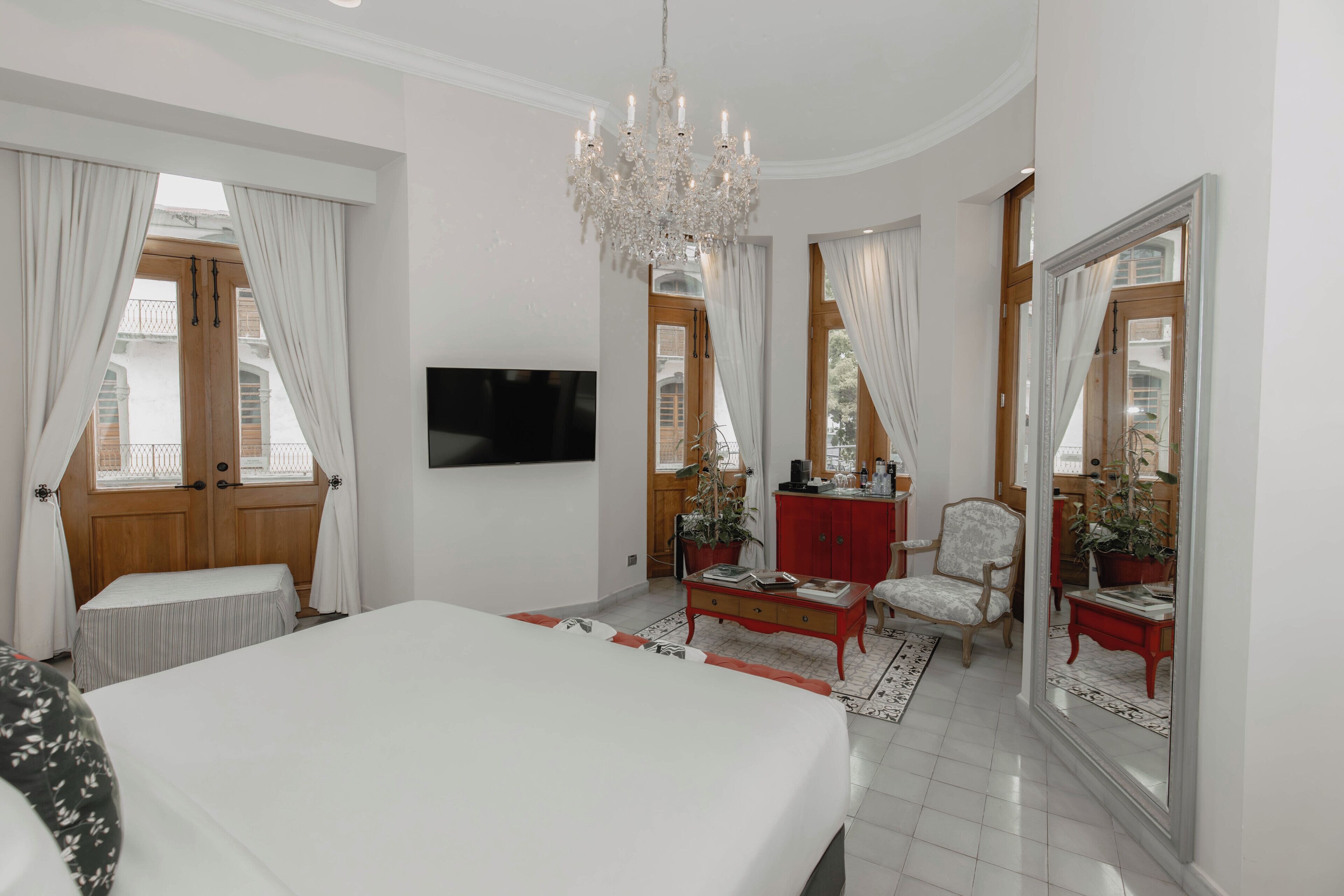 A bedroom with a large bed, a TV mounted on the wall, a chandelier, and a red side table with a chair.