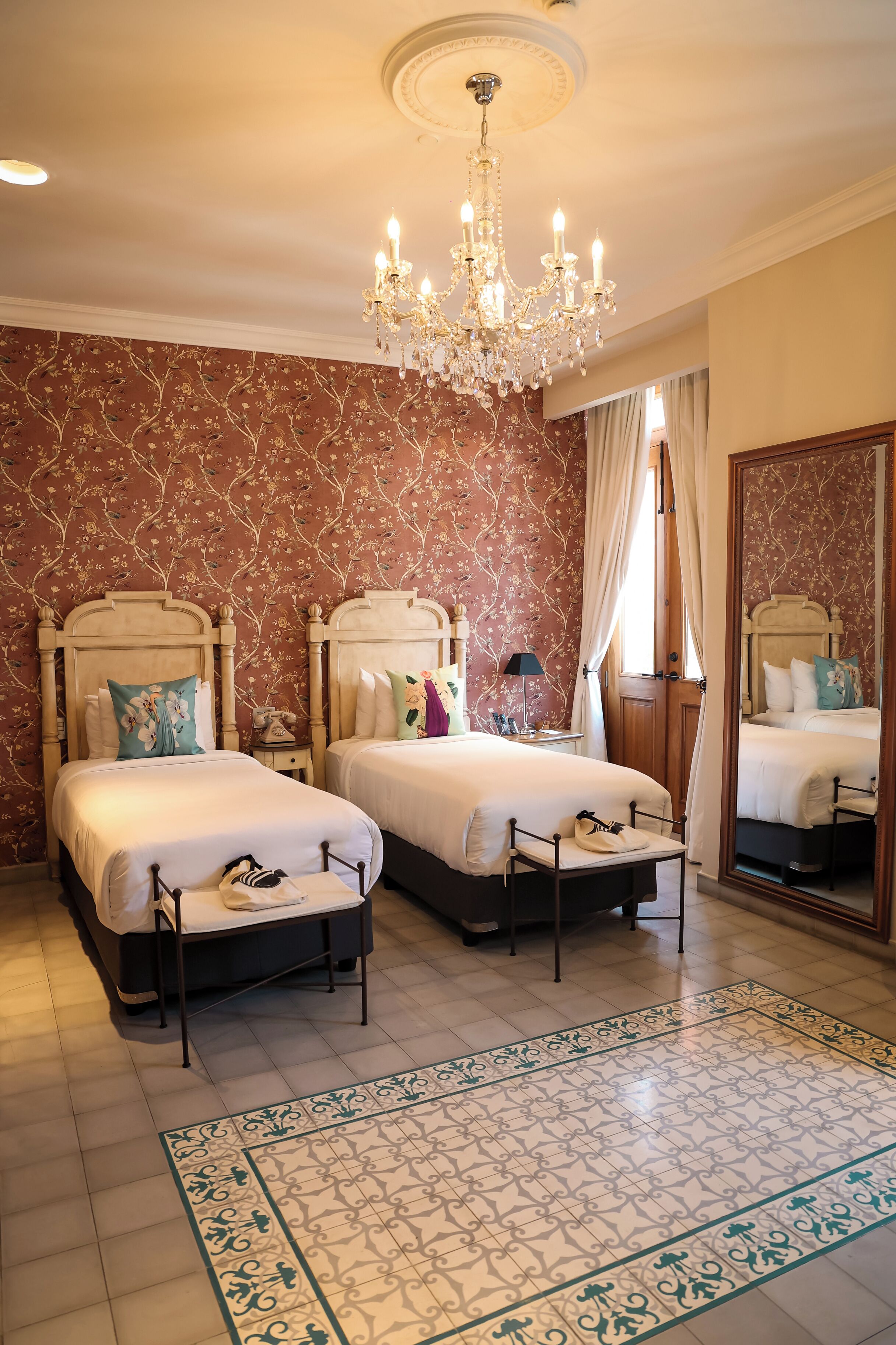 Une chambre d’hôtel avec deux lits, un lustre, un papier peint à motifs floraux et un grand miroir.
