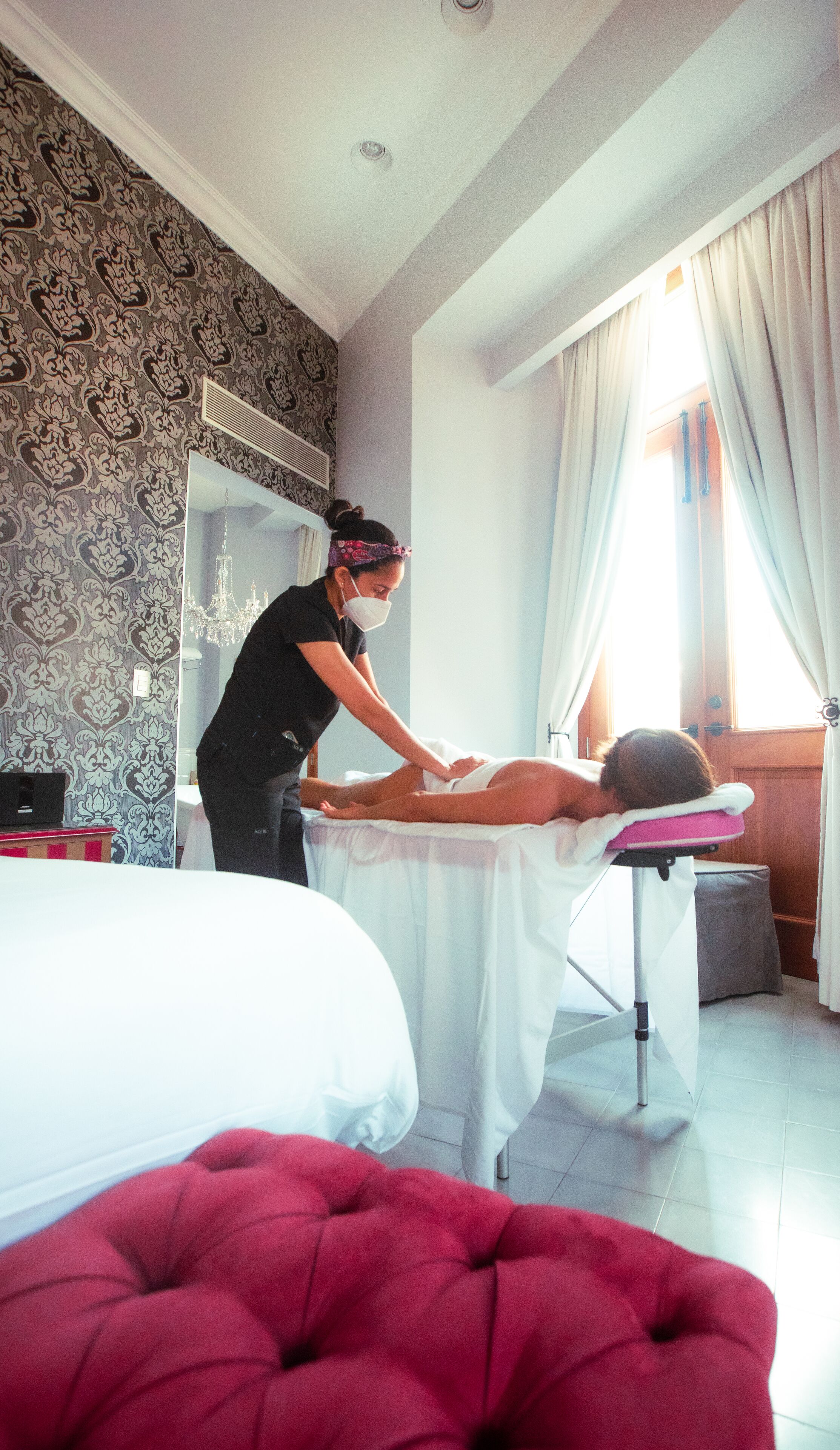 A massage therapy room with a bed, a massage table, and a red ottoman.