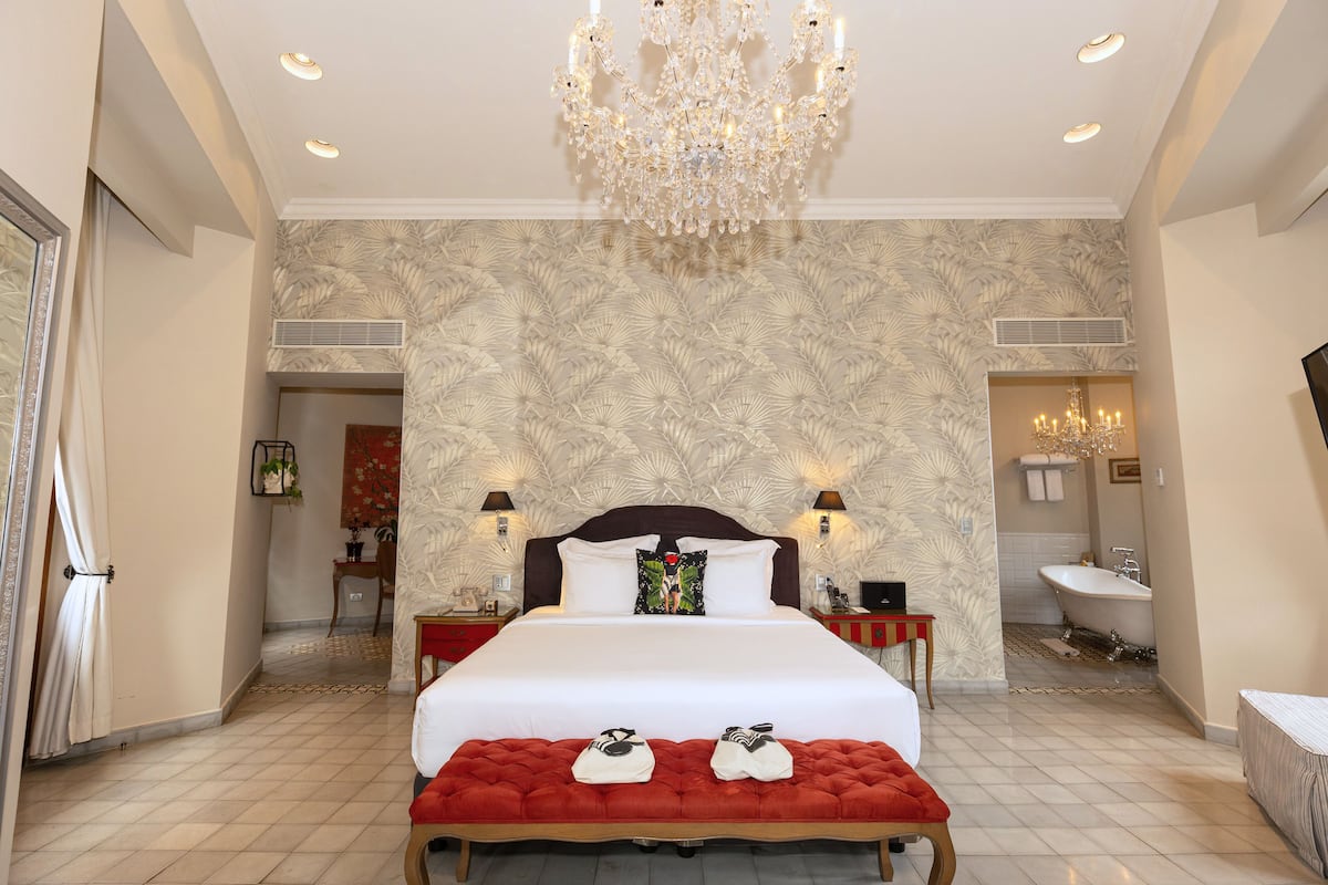 A spacious bedroom with a large bed, a red bench, a chandelier, and a bathroom with a bathtub.