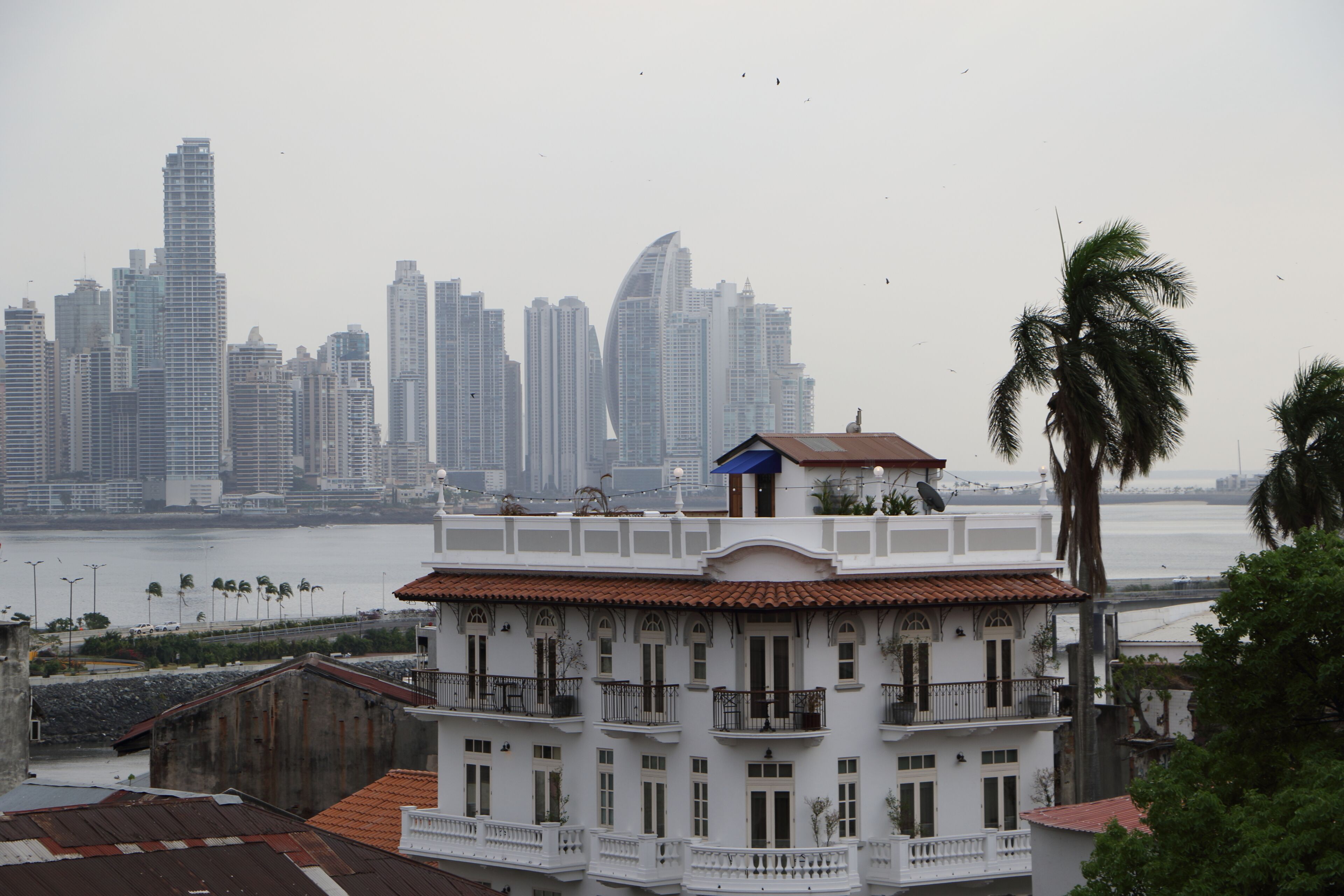 A white building with balconies overlooking a body of water and a city skyline in the background.