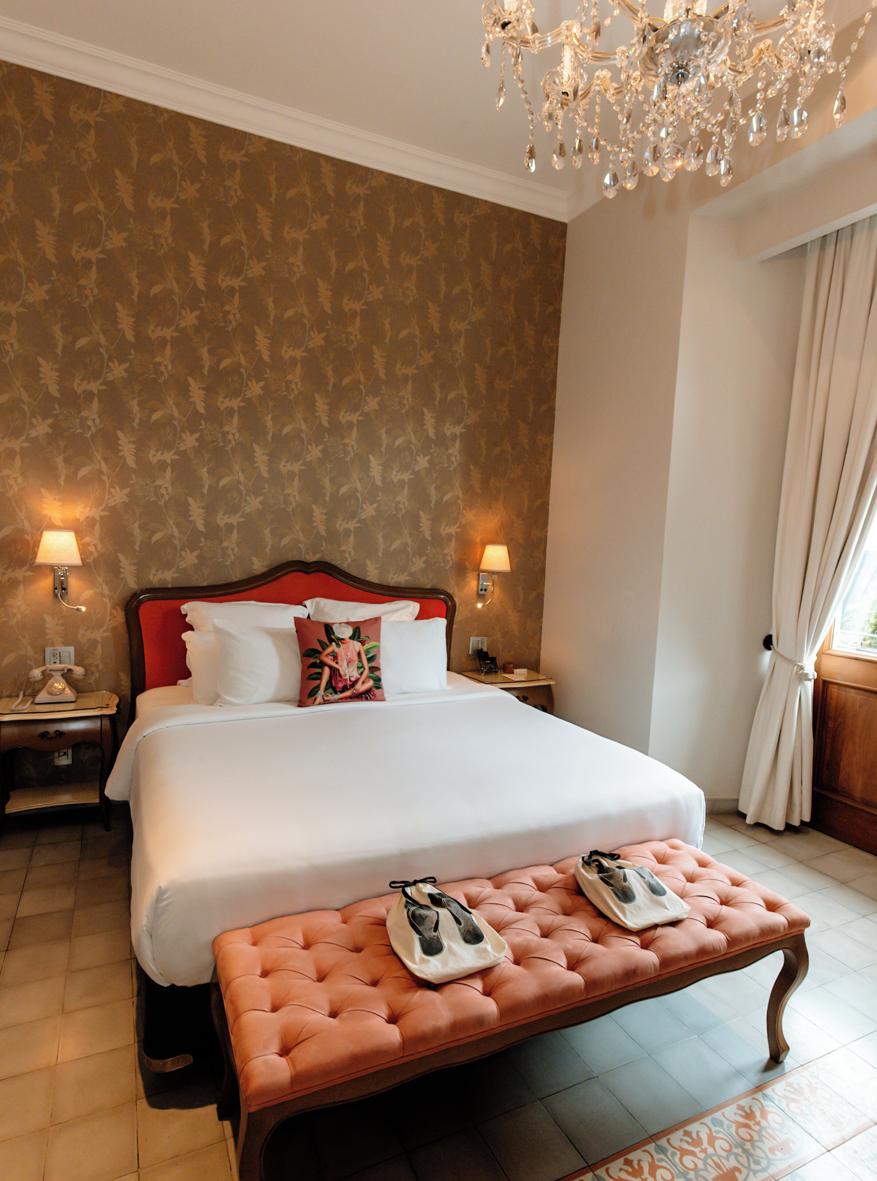 A hotel room with a large bed, a bench with slippers, two bedside lamps, a chandelier, and a patterned wallpaper.