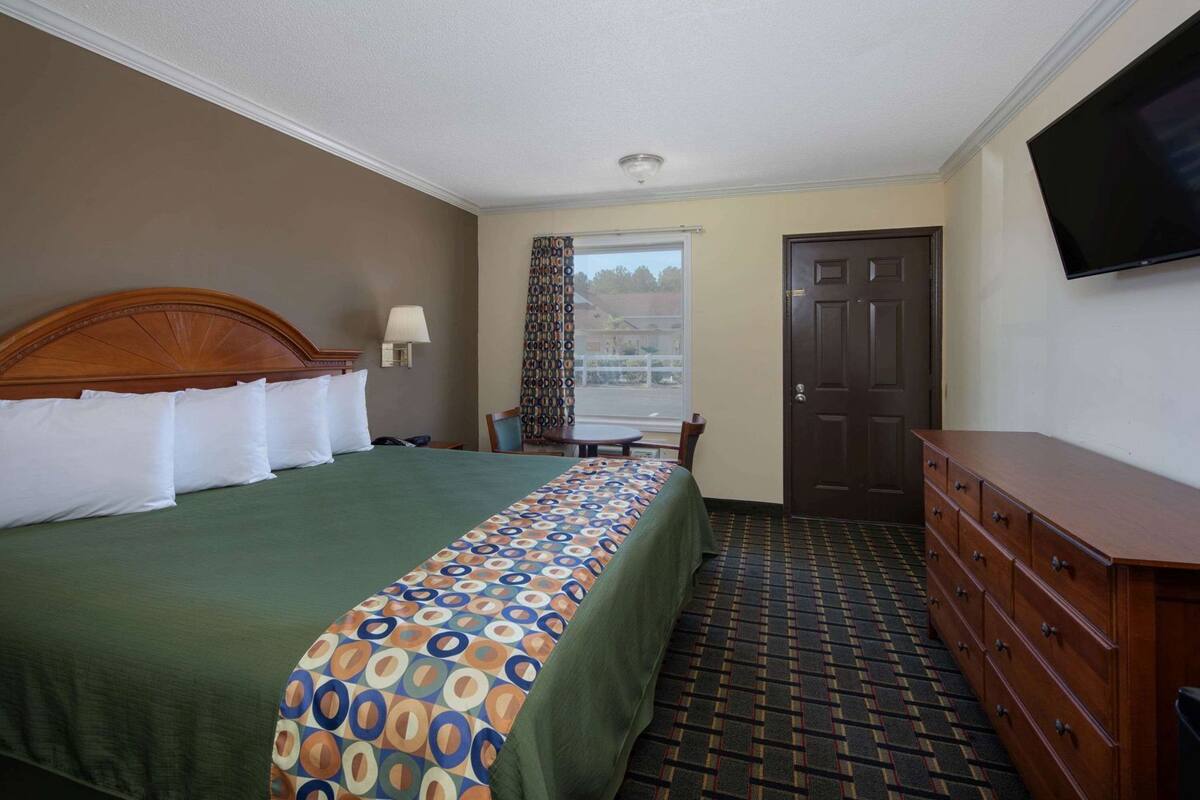 A hotel room with a bed, a dresser, a television, a window with curtains, and a door.