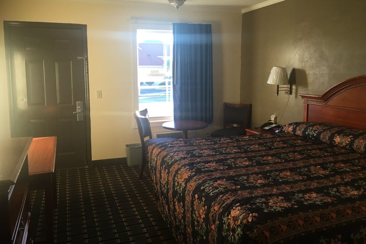 A hotel room with a bed, a desk, a chair, a small table, a lamp, and a window with curtains.