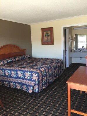 A hotel room with a bed, a wooden headboard, a floral-patterned bedspread, a small table, a mirror, and a door leading to a bathroom.