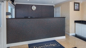 Uma área de recepção com um balcão escuro, um relógio na parede e um tapete de entrada com a inscrição "Travelodge".