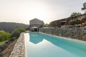 Una piscina de paredes de piedra con superficie de agua azul cristalina, rodeada de abundante vegetación y un edificio de piedra al fondo.