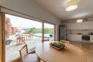 Una cocina moderna con armarios blancos, un comedor con mesa redonda y vistas a una piscina y jardín a través de amplios ventanales.