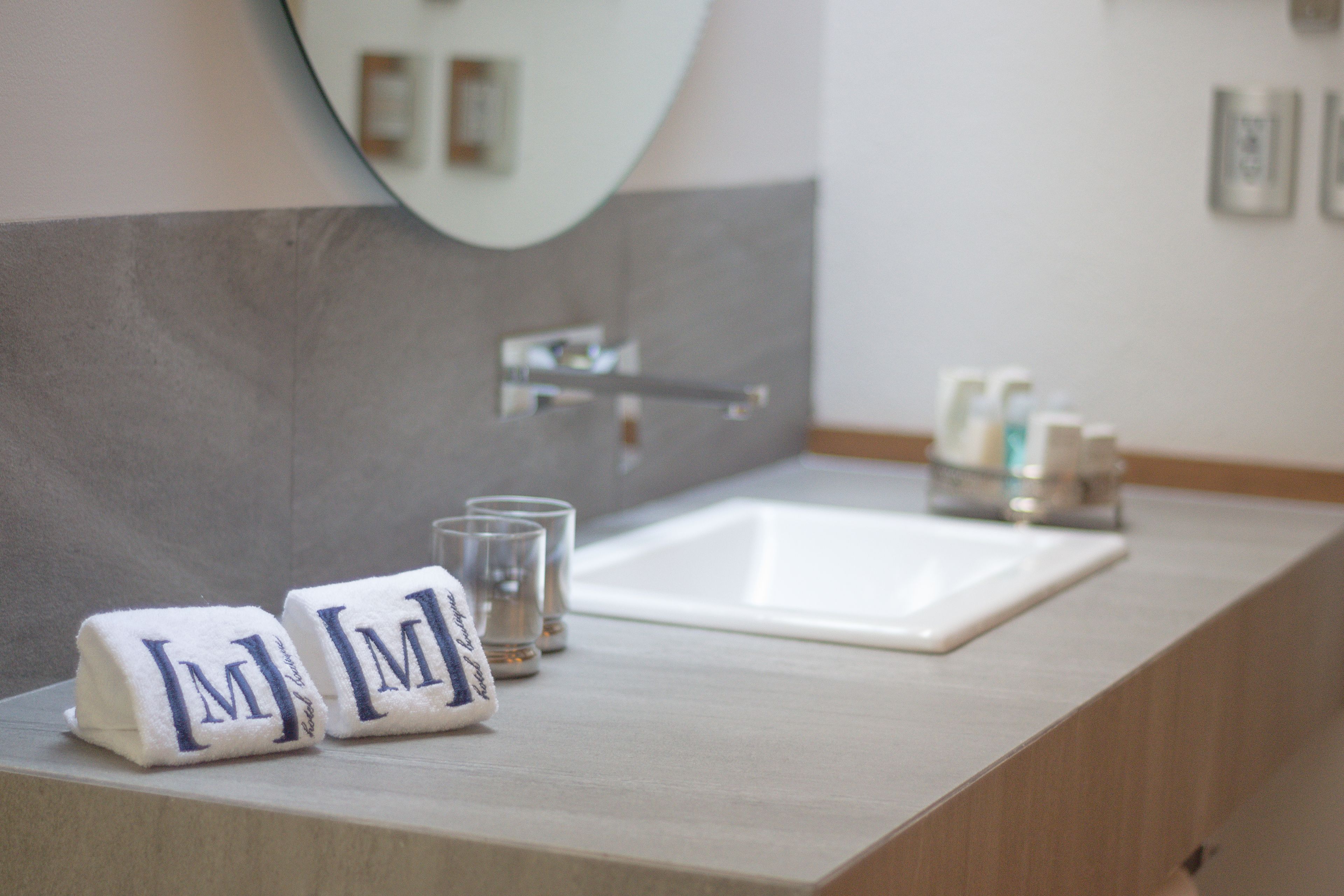 Une salle de bain avec un lavabo blanc, deux serviettes ornées des initiales « M », et un miroir au-dessus du lavabo.