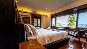 Quarto moderno com uma cama grande, mesa de cabeceira de madeira e vista para a cidade através de janelas amplas.