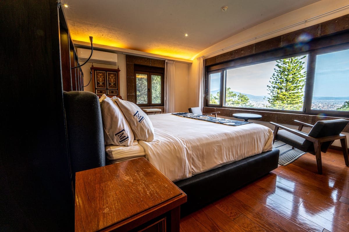 Ein modernes Schlafzimmer mit einem großen Bett, einem Nachttisch aus Holz und Blick auf eine Stadtlandschaft durch große Fenster.