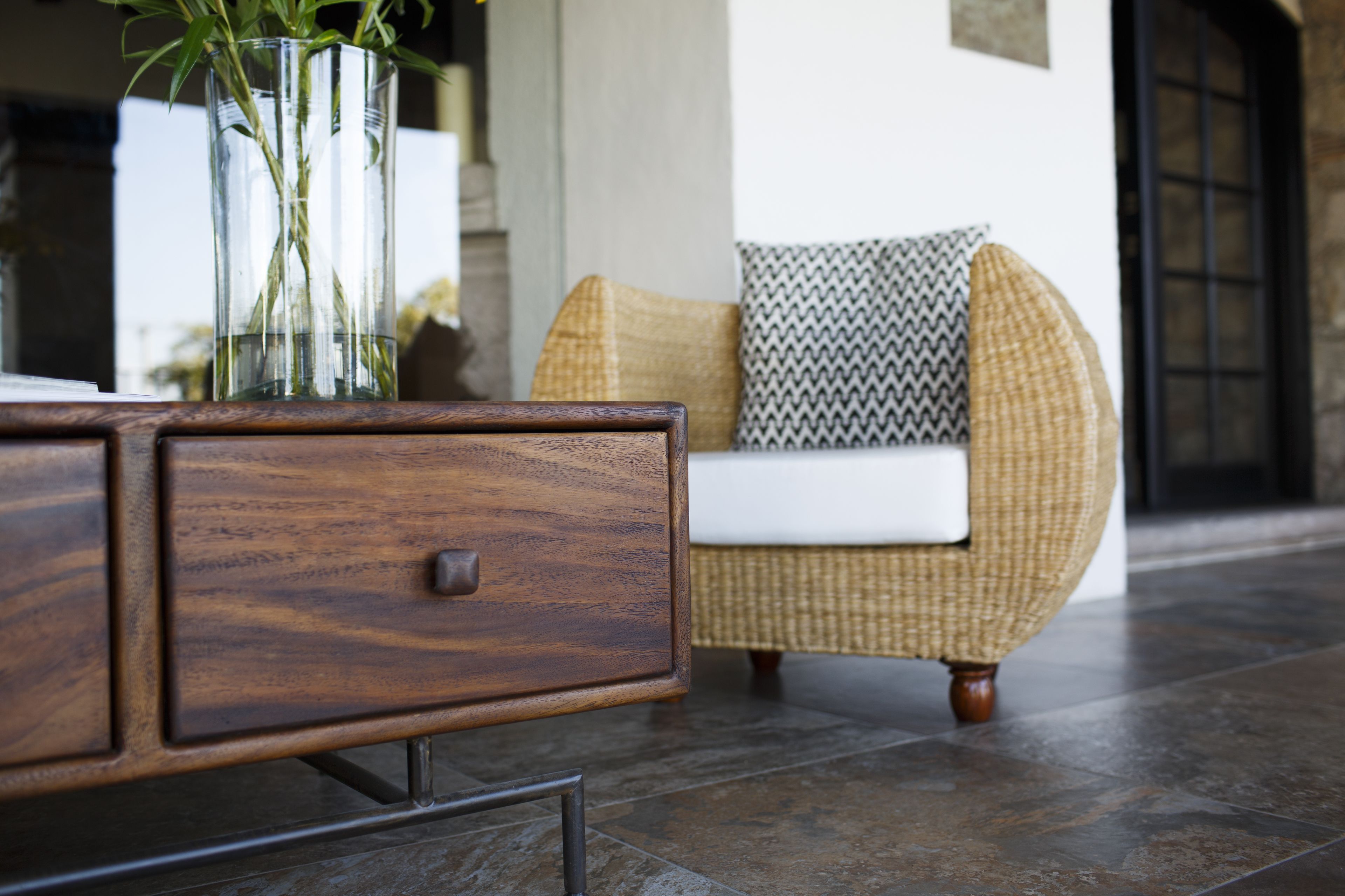 Un mueble de madera con un cajón, una silla de mimbre con un cojín estampado y un florero de vidrio con plantas verdes.