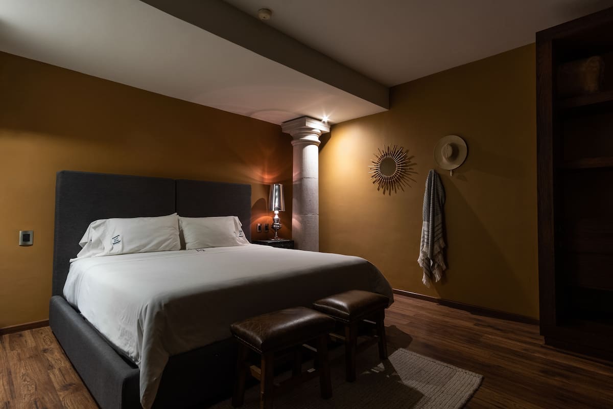 Ein ordentlich bezogenes Bett mit Kopfteil, ein Nachttisch mit Lampe, eine Bank und eine Wanddekoration in Form einer Sonne.