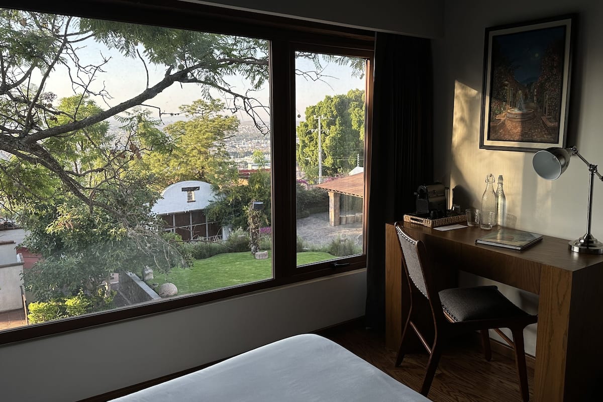 Ein Zimmer mit großem Fenster, Schreibtisch mit Stuhl, Lampe und einem gerahmten Bild an der Wand.