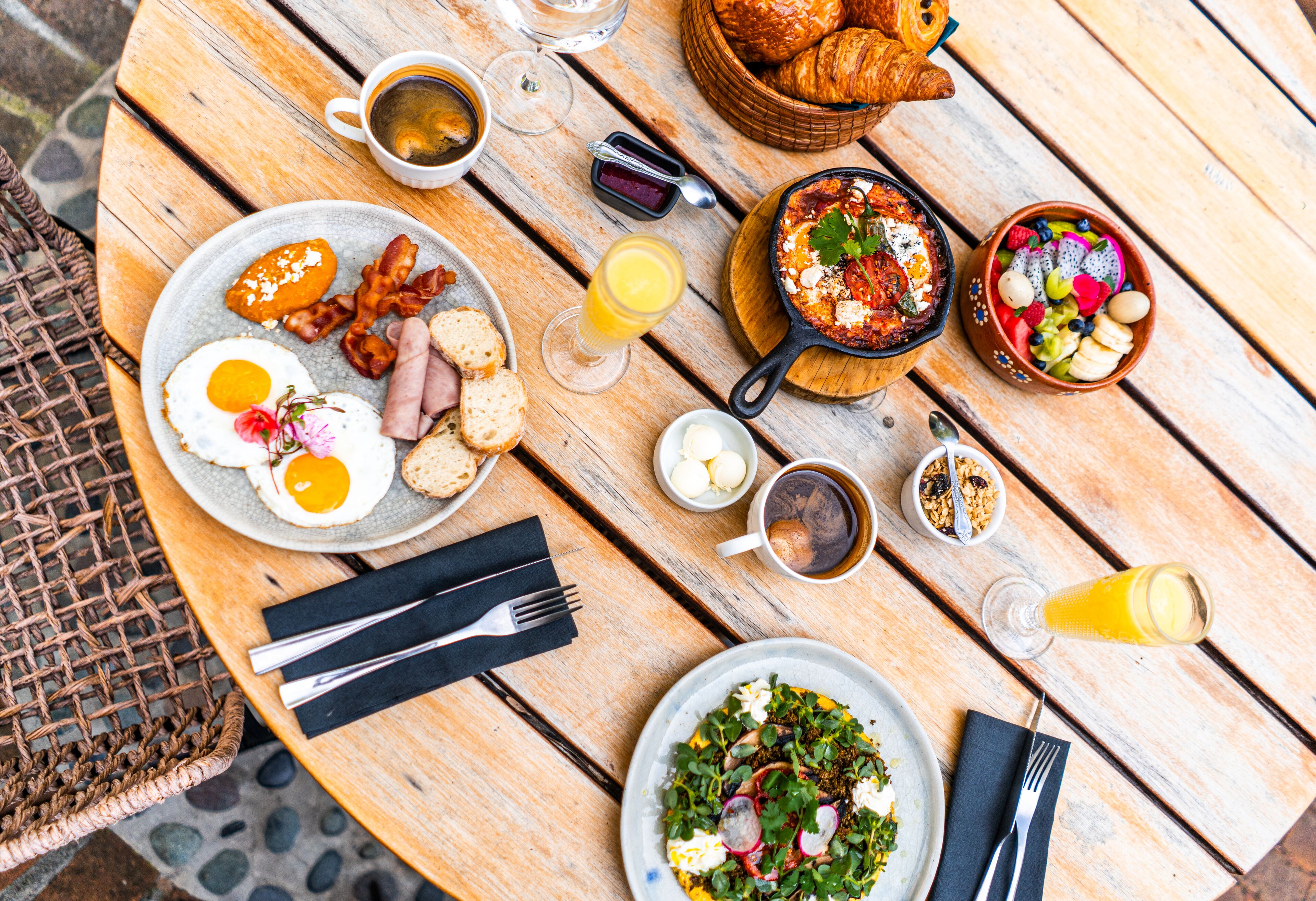 A wooden table with breakfast items including eggs, bacon, toast, a bowl of fruit, and a skillet dish.