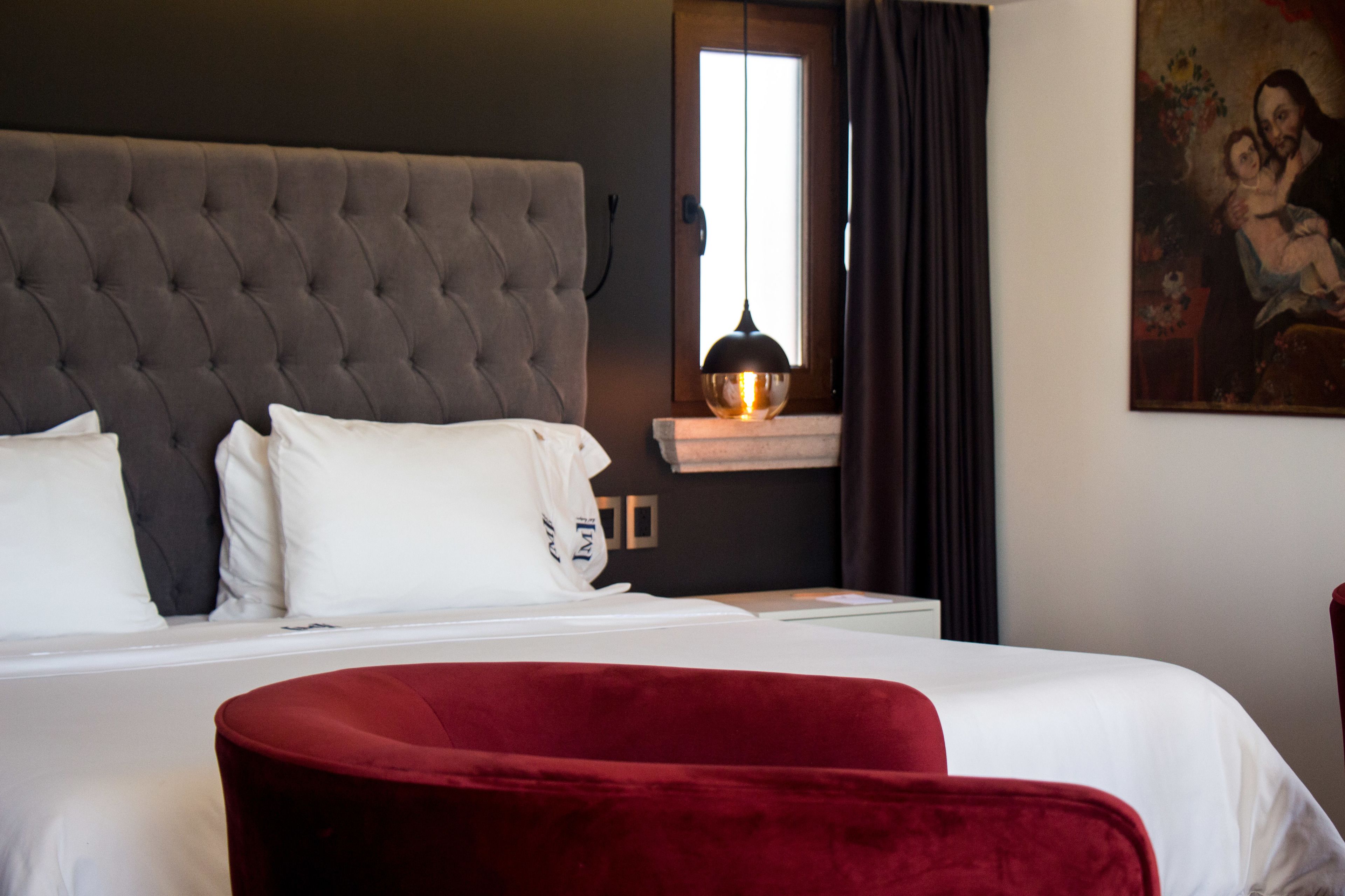 Ein Hotelzimmer mit einem großen Bett, einem roten Sessel, einem Bild an der Wand und einem Fenster mit Vorhängen.