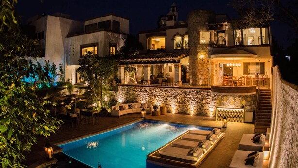 Uma área de piscina luxuosa, com parede de pedra, espreguiçadeiras e um bar.