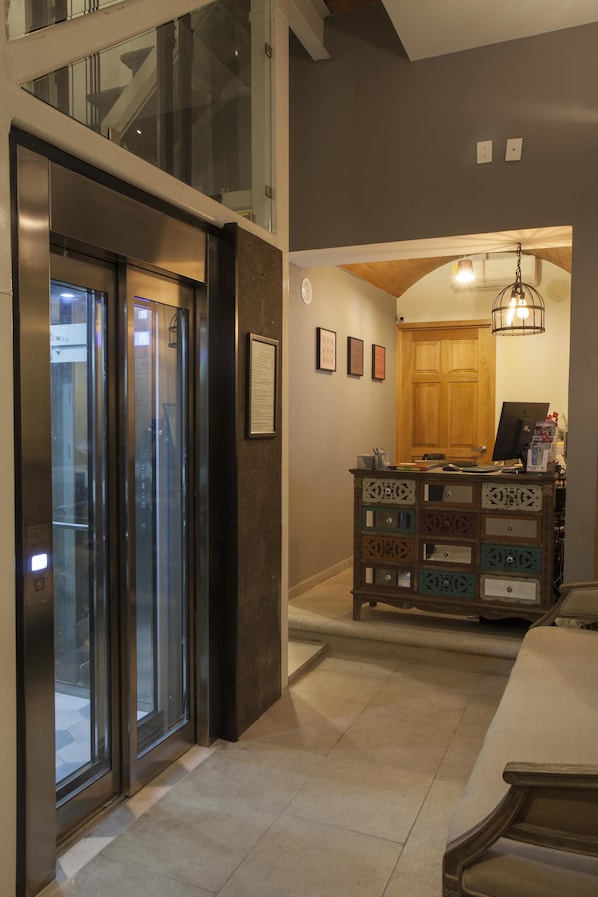 A modern interior with a glass elevator, a wooden door, and a decorative cabinet. - Casa Rosa Gran Hotel Boutique (Puebla)
