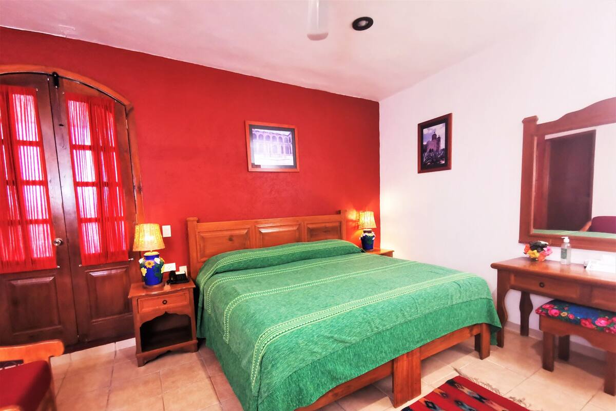 Una habitación con cama de madera, paredes rojas, armario de madera, mesita de noche con lámpara y espejo.