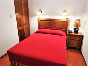 Une chambre à coucher avec une tête de lit en bois, une couvre-lit rouge, deux lampes fixées au mur, une table de chevet avec une lampe et un téléphone.