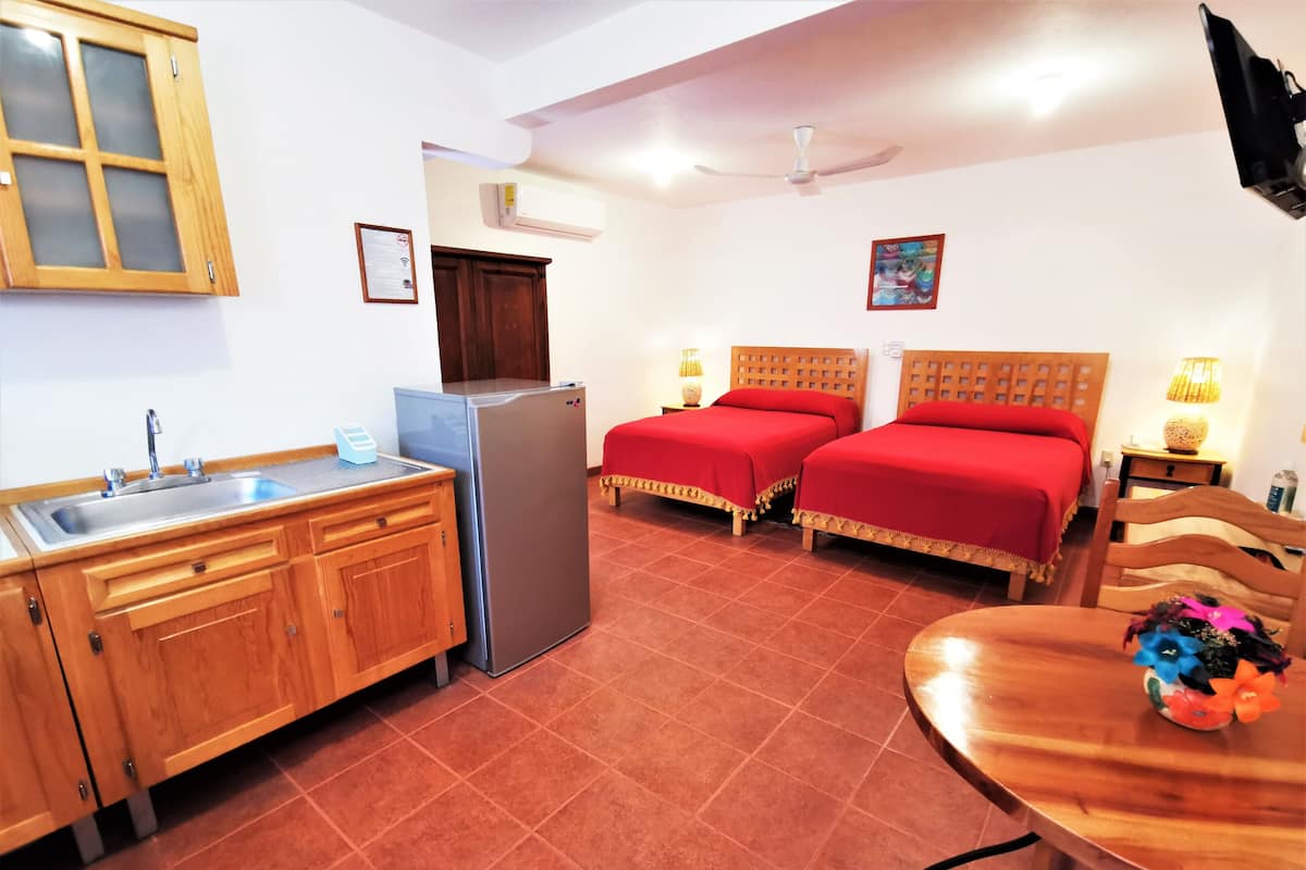 Una habitación de hotel compacta con kitchenette, dos camas individuales, una mesa de comedor de madera y un ventilador de techo.