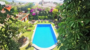 Une piscine entourée d’une végétation luxuriante, avec une vue sur un paysage urbain où l’on aperçoit des bâtiments et des montagnes au loin.