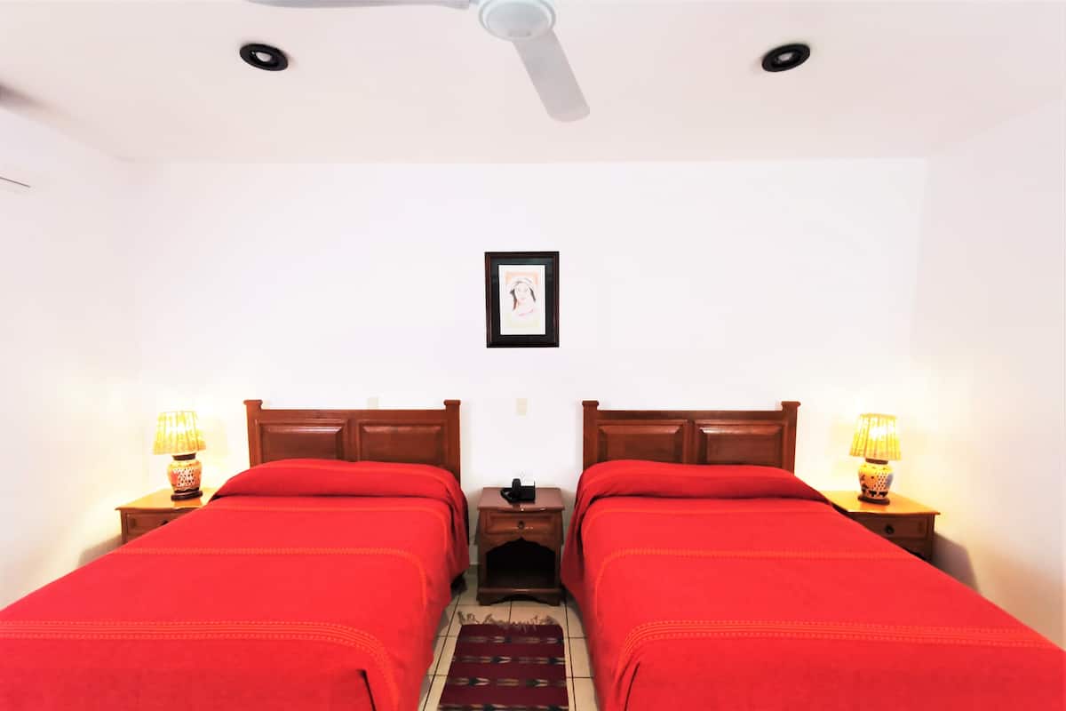 Una habitación con dos camas individuales, cada una con colcha roja y cabecero de madera. Hay dos mesitas de noche con lámparas, un ventilador de techo y un cuadro enmarcado en la pared.