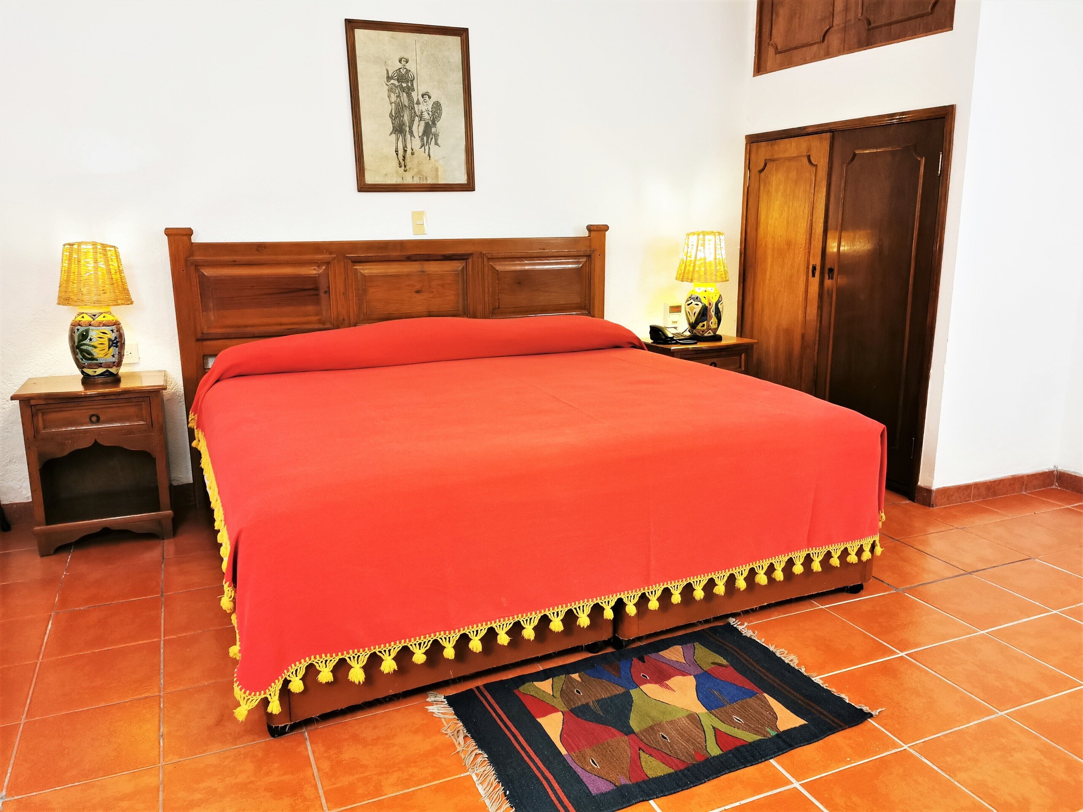 A neatly made bed with a red blanket, a wooden headboard, a small bedside table with a lamp, and a colorful rug on a tiled floor.
