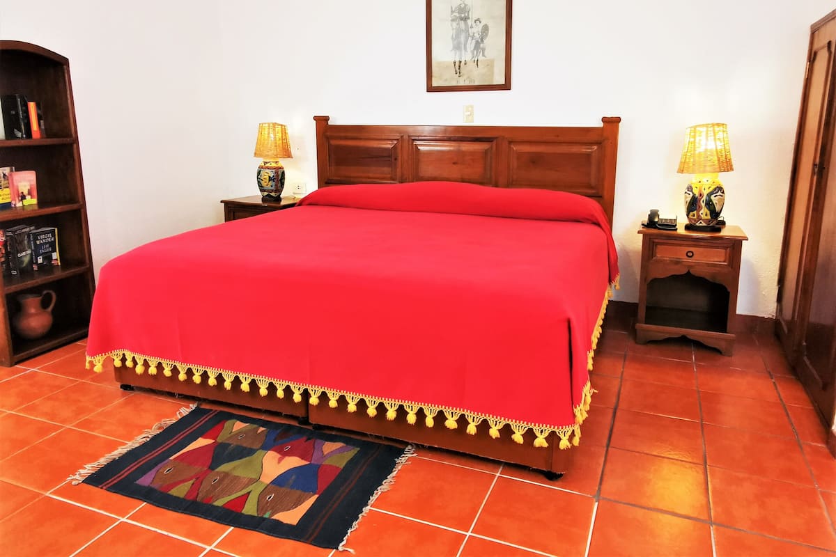 Una cama bien hecha con una colcha roja y un borde decorativo, flanqueada por dos mesitas de noche con lámparas, una estantería y un cuadro en la pared.
