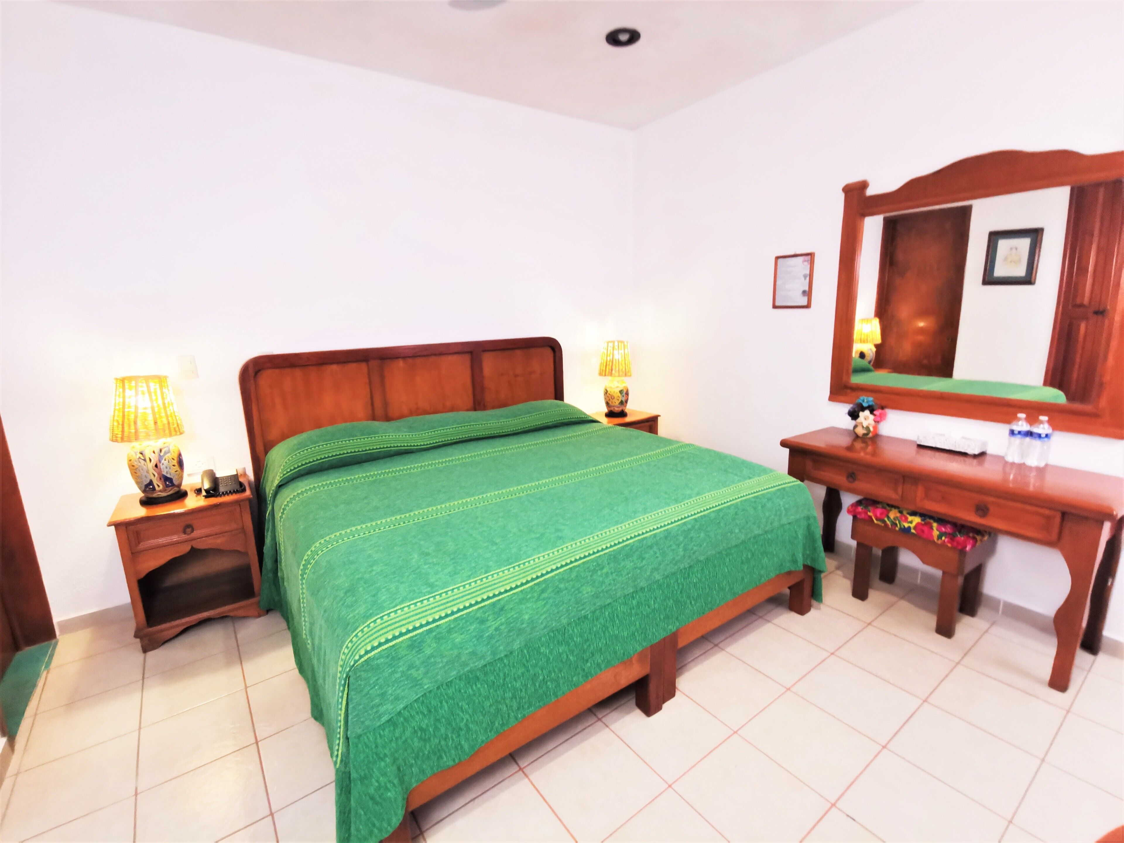 A bedroom with a wooden bed, green bedspread, two bedside tables with lamps, a wooden dresser, and a mirror.