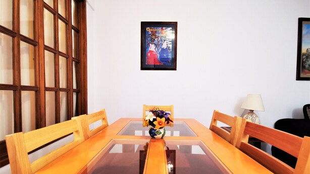 A dining table with a glass top, surrounded by wooden chairs, in a room with a large window and a framed picture on the wall.