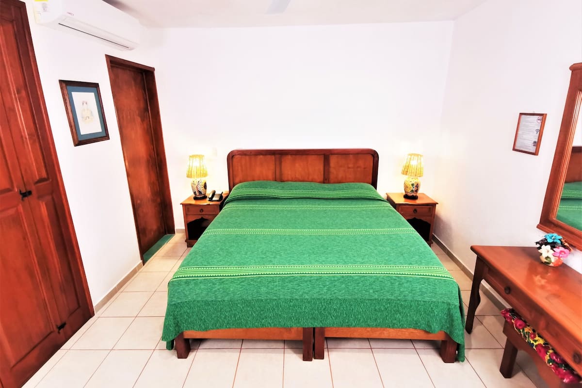 Un dormitorio con cama de madera, colcha verde, dos mesitas de noche con lámparas, un ventilador de techo y un armario de madera.
