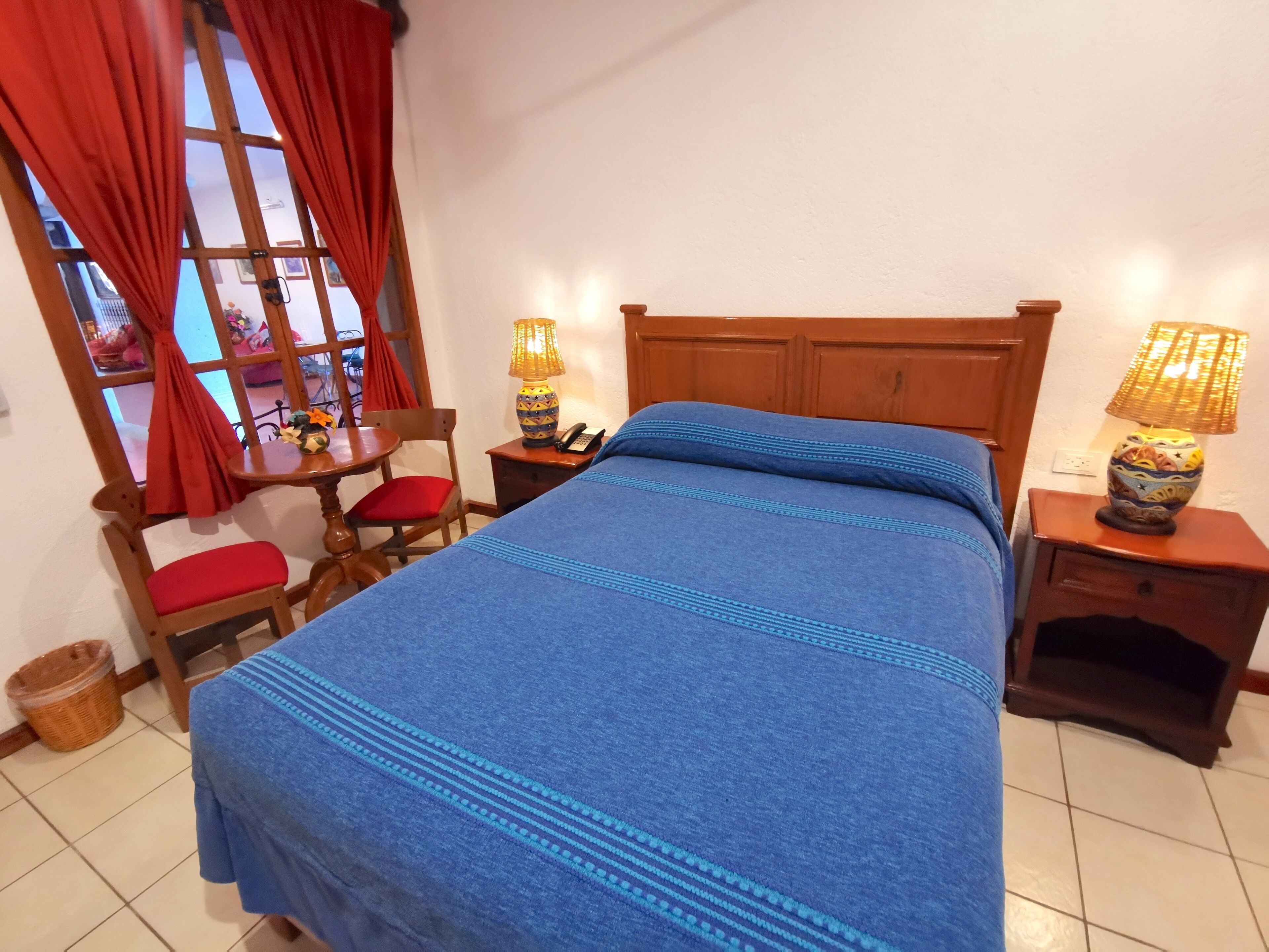 A bedroom with a wooden headboard, a blue bedspread, a chair with a red cushion, a table with a lamp, and a window with red curtains.