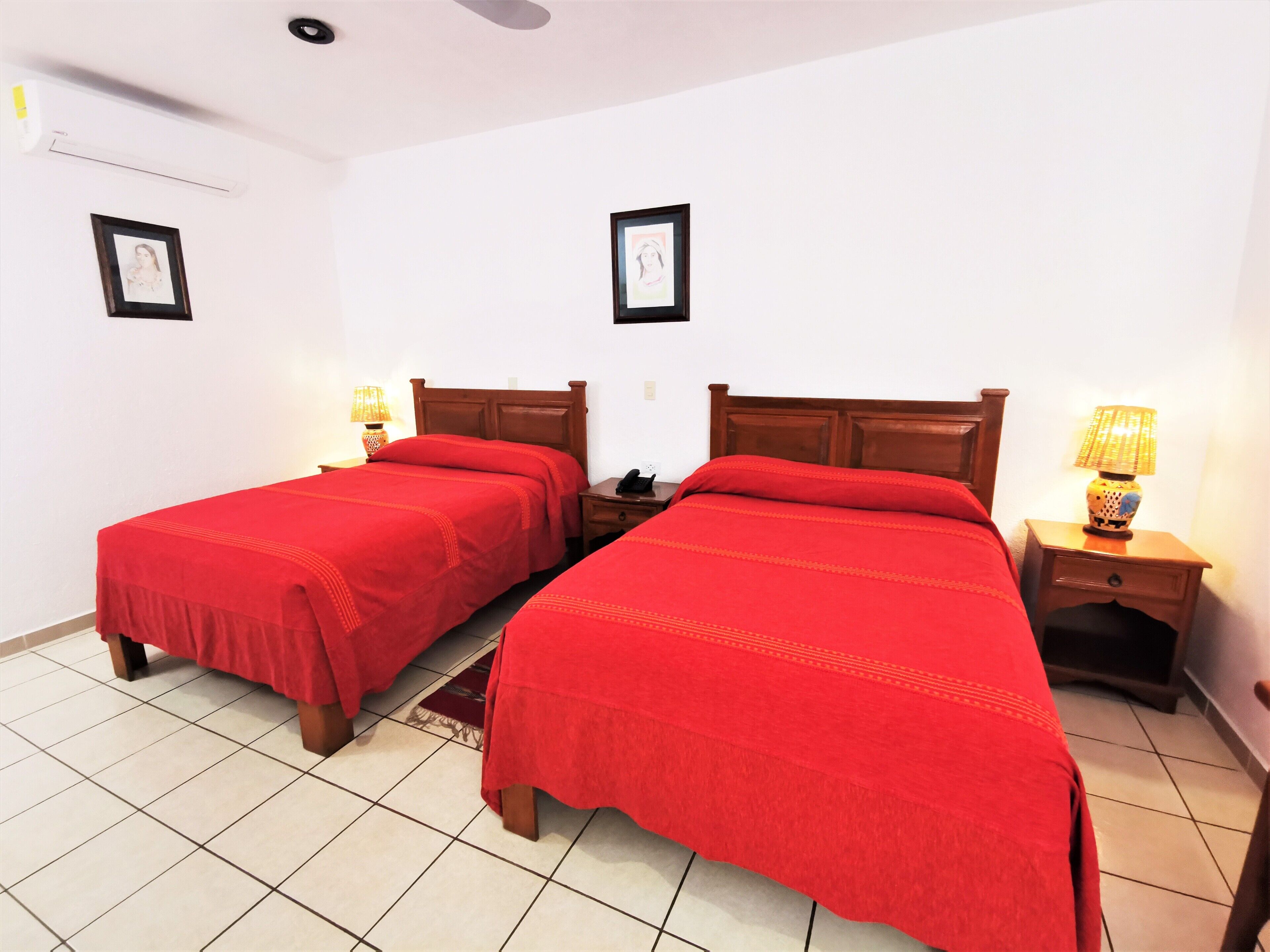 Une chambre d’hôtel avec deux lits simples, chacun recouvert d’une couverture rouge et doté d’une tête de lit en bois. On y trouve deux tables de chevet avec des lampes, et les murs sont blancs, tandis que le sol est carrelé.