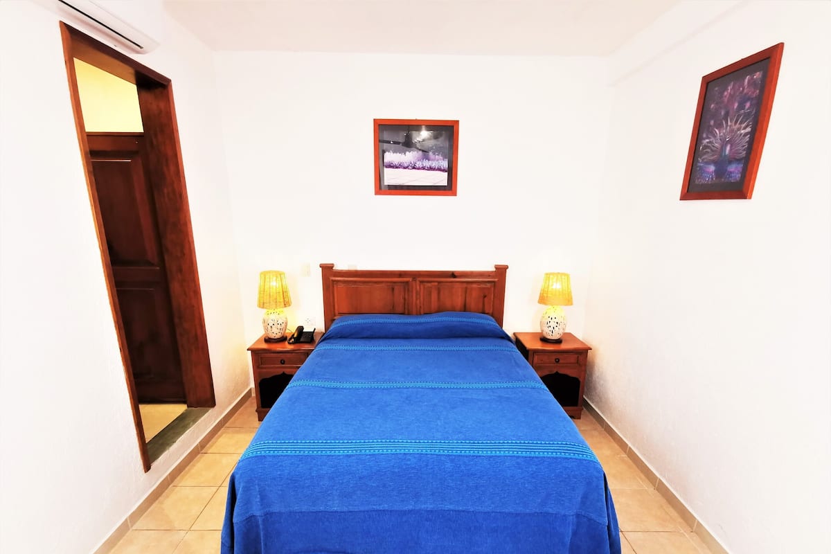 Un dormitorio con una cama de madera, dos mesitas de noche con lámparas, un cuadro enmarcado en la pared y una puerta a la izquierda.