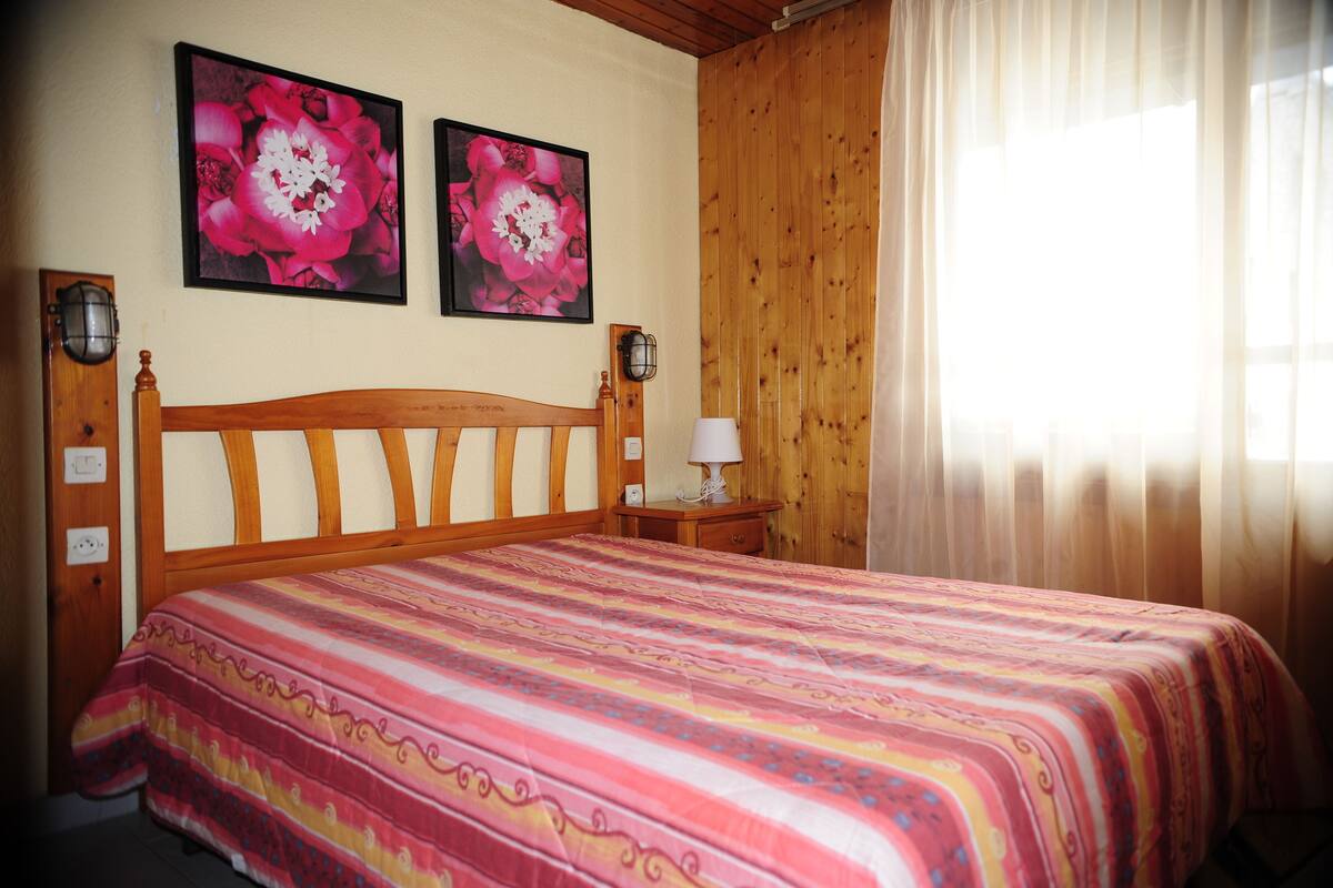 Une chambre à coucher avec un lit en bois, deux lampes fixées au mur et une fenêtre ornée de rideaux légers.