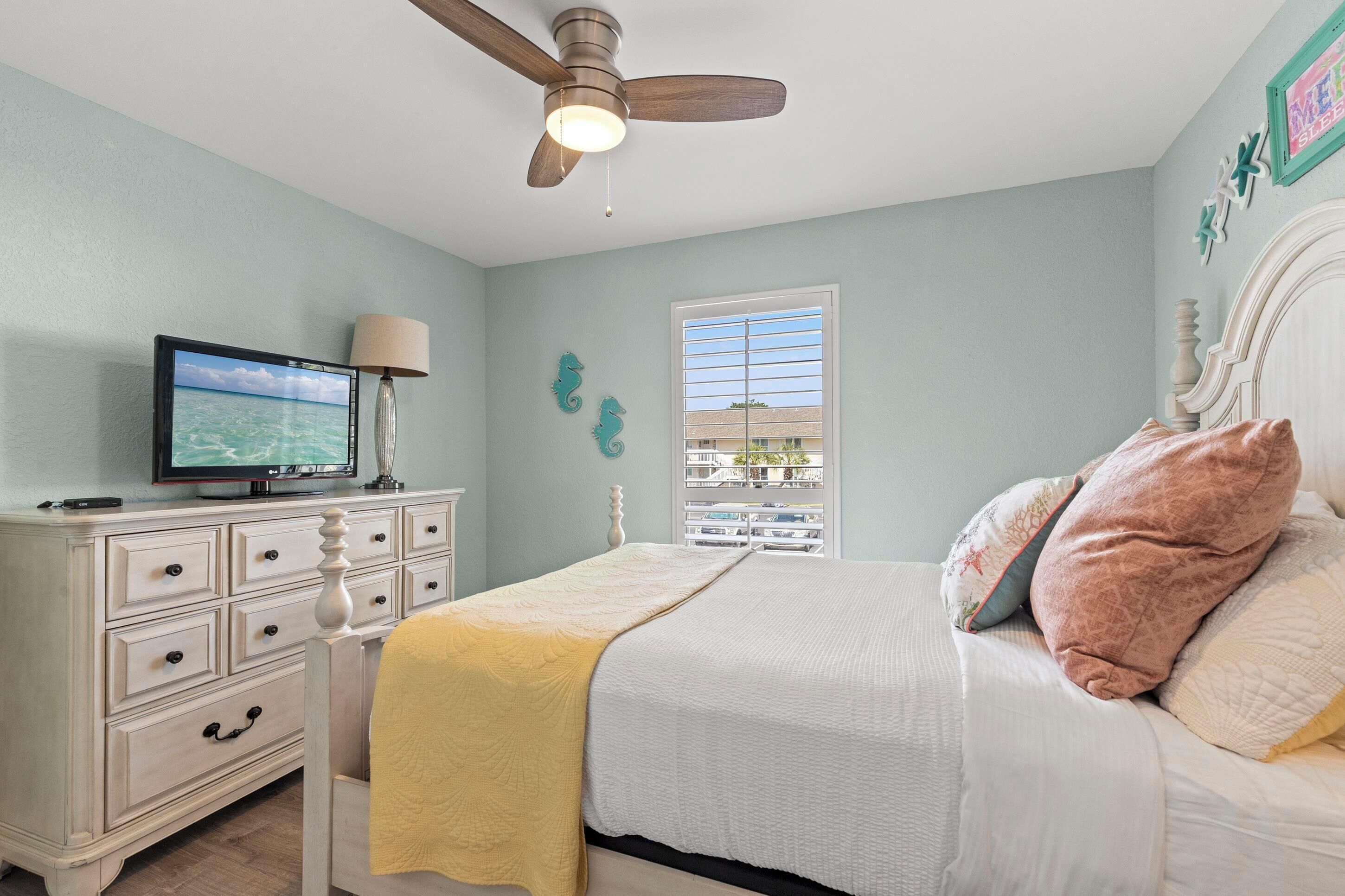 A bedroom with a bed, a dresser, a television, a ceiling fan, and a window with a view.