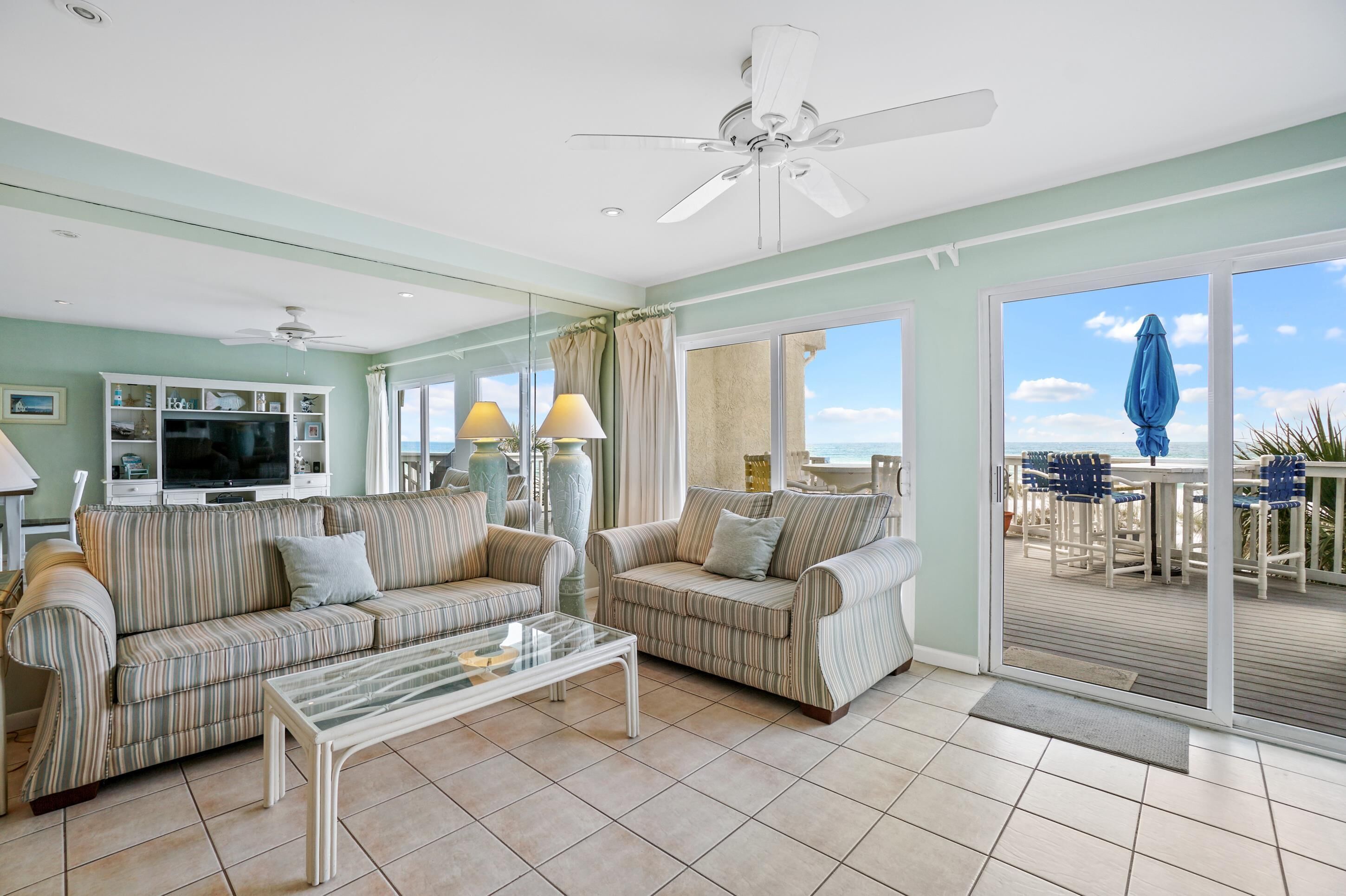 A living room with striped sofas, a glass coffee table, ceiling fans, and a view of the ocean through large sliding glass doors.
